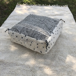 May include: A black and white patterned floor cushion with a white fringe. The cushion is sitting on a white rug.