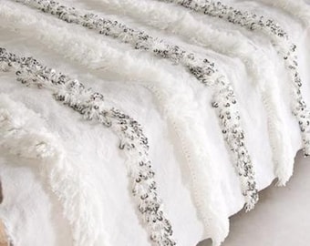 May include: White throw blanket with a fringe border and silver accents. The blanket is draped over a bed.