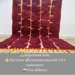 May include: A burgundy rug with a geometric pattern of light tan rectangles and lines. The rug has a thick, textured pile and a fringe border with orange and cream accents. The rug is displayed on a light-colored surface.