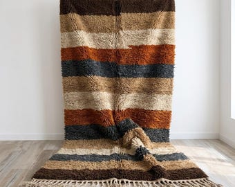 Op de afbeelding: Een rechthoekig hoogpolig vloerkleed met horizontale strepen in tinten donkerbruin, beige, crème, roestoranje en antracietgrijs. Het kleed heeft een dikke, getextureerde pool en is afgewerkt met geknoopte franjes aan de onderkant.
