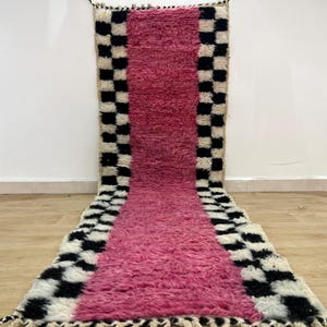 Op de afbeelding: Een lang roze en zwart tapijt met een geruit patroon. Het tapijt is gemaakt van wol en heeft een zachte, pluche textuur.