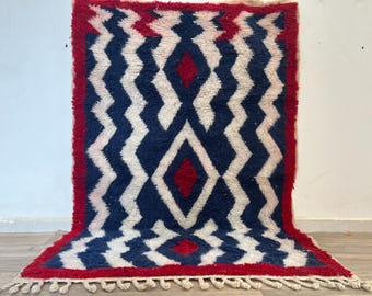 Puede incluir: Una alfombra rectangular con un patrón geométrico. La alfombra presenta un fondo azul marino con líneas en zigzag blancas y formas de diamante rojas. El borde es rojo y la alfombra tiene una franja en el borde inferior.