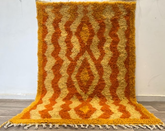 May include: A rectangular, shaggy rug with a geometric pattern in shades of orange and yellow. The design features zig-zag lines and diamond shapes. The rug has a fringe along the bottom edge.