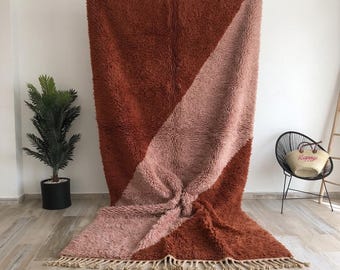 Op de afbeelding: Een groot, rechthoekig hoogpolig vloerkleed met een diagonaal kleurblokontwerp in diep terracotta en zacht oudroze. Het kleed heeft een dikke, getextureerde pool en is afgewerkt met een rij geknoopte franjes aan de onderzijde.