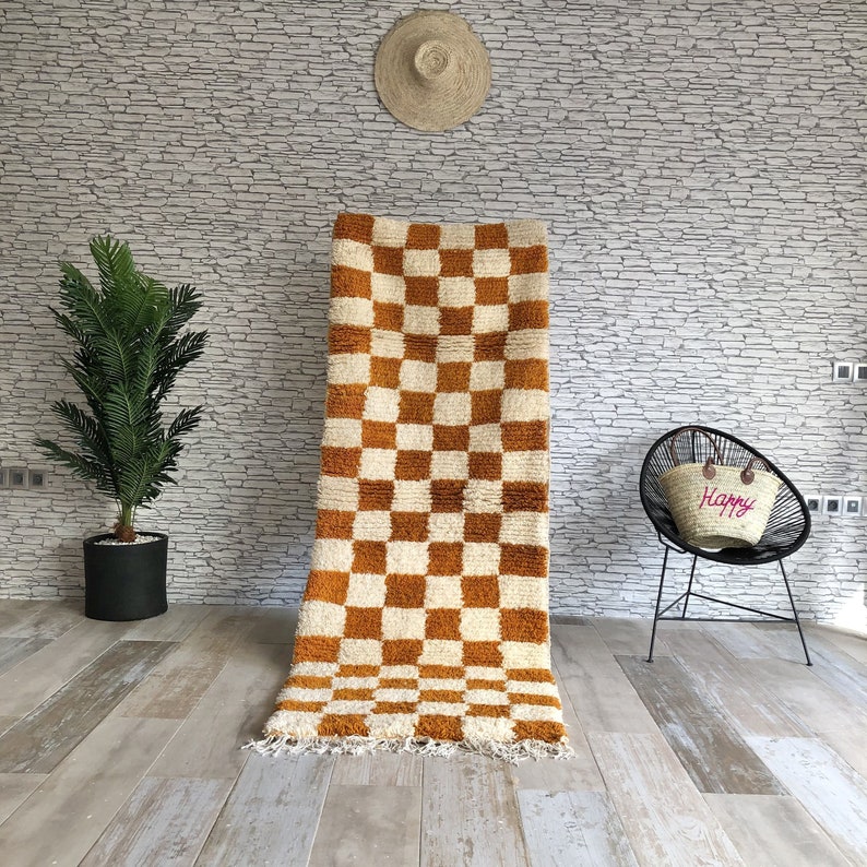 May include: A brown and white checkered rug with a fringe border. The rug is hanging on a wall in a room with a white brick wall, a potted plant, and a chair with a straw bag.