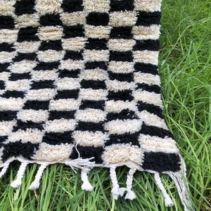 May include: Black and white checkered rug with a fringe border. The rug is made of a soft, fluffy material and has a geometric pattern.