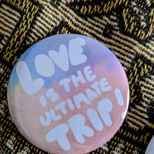 K&ouml;nnte beinhalten: Runder Button mit pastellfarbenem Farbverlauf in Blau-, Rosa- und Pfirsicht&ouml;nen. Der wei&szlig;e Text lautet "LOVE IS THE ULTIMATE TRIP!". Der Button liegt auf einem gelb-schwarz gemusterten Stoff.