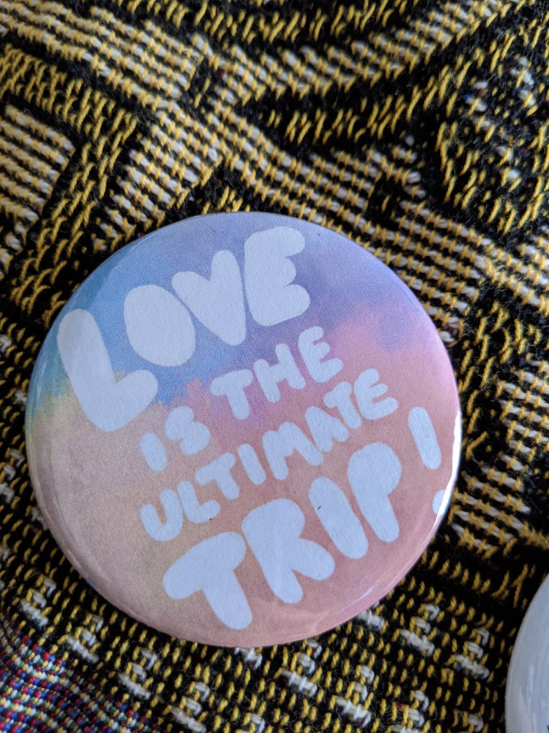 K&ouml;nnte beinhalten: Runder Button mit pastellfarbenem Farbverlauf in Blau-, Rosa- und Pfirsicht&ouml;nen. Der wei&szlig;e Text lautet "LOVE IS THE ULTIMATE TRIP!". Der Button liegt auf einem gelb-schwarz gemusterten Stoff.