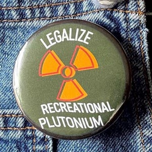 Puede incluir: Un botón redondo verde oliva con las palabras "LEGALIZE RECREATIONAL PLUTONIUM" en texto blanco. El botón presenta un símbolo de radiación amarillo y naranja. El botón está prendido a una chaqueta vaquera.