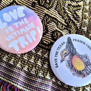 K&ouml;nnte beinhalten: Zwei runde Ansteckbuttons. Einer zeigt den Text "LOVE IS THE ULTIMATE TRIP!" in Wei&szlig; auf pastellfarbenem Hintergrund. Der andere Button zeigt eine detaillierte Illustration eines Pr&auml;riehuhns mit dem Text "SAVE THE TEXAS PRAIRIE CHICKEN".