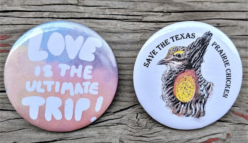 K&ouml;nnte beinhalten: Zwei runde Buttons. Einer hat einen bunten Hintergrund mit den wei&szlig;en Worten "LOVE IS THE ULTIMATE TRIP!". Der andere Button zeigt eine Zeichnung eines Pr&auml;riehuhns mit dem Text "SAVE THE TEXAS PRAIRIE CHICKEN" in Schwarz.