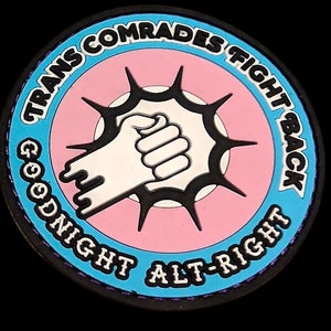以下が含まれることがあります： 縁に「Trans Comrades Fight Back」と「Goodnight Alt-Right」の文字が書かれた青とピンクの円形のワッペン。ワッペンの真ん中には、拳を突き合わせた2つの手の白黒の画像があります。