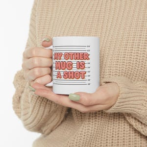 May include: White ceramic mug with the text "MY OTHER MUG IS A SHOT" in red and orange lettering. The mug has a height chart design on the side, with measurements in inches.