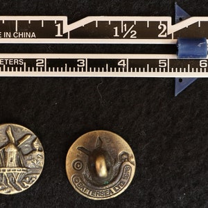 May include: Two antique brass buttons with detailed designs. One features a windmill scene, the other a stylized boat and the text "BATTERSEA LTD 1976". A ruler shows measurements in inches and centimeters.