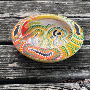 May include: A hand-painted, round ceramic dish with a vibrant dot-painted design. The colours include yellow, orange, green, red, black, and white. The dish is set against a weathered wooden surface.