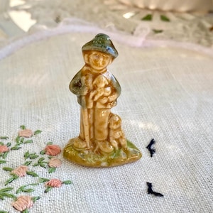 Può includere: Piccola statuetta in ceramica smaltata raffigurante un pastore con un bastone, due pecore e un cane. Il pastore indossa un cappello e un cappotto verdi. La statuetta è su una base verde.