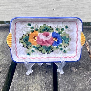 May include: A small, rectangular ceramic dish with a blue and white border. The dish is decorated with a hand-painted floral design featuring pink, yellow, blue, and green flowers.