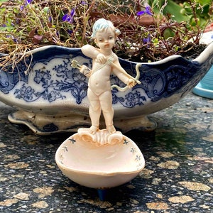 Könnte beinhalten: Eine Porzellanfigur eines Cherubs, der einen Bogen und Pfeil hält, steht auf einer rosa-weißen, muschelförmigen Schale. Der Cherub ist in einem hell elfenbeinfarbenen Ton mit blauen Akzenten bemalt.