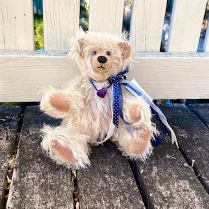 以下が含まれることがあります： ダークブラウンの鼻と目、薄茶色の足と耳を持つ、クリーム色のテディベア。 白い水玉模様の青いリボンと紫色のハートペンダントが付いています。 熊は風化した木製の表面に座っています。