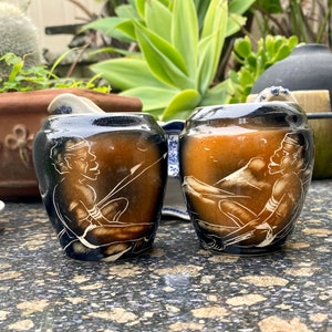 May include: Two small ceramic jars with brown and black glaze. Each jar features a white design of a person in a seated position.