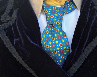 May include: A blue tie with a geometric pattern of red, yellow, and green squares on a white background. The tie is knotted and worn with a dark blue velvet jacket.