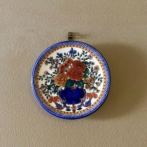 May include: Round ceramic decorative plate with a floral design. The plate features a blue rim with a white and gold border. The center of the plate has a blue vase filled with orange and red flowers.
