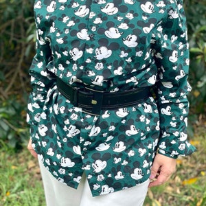 May include: A green button-down shirt with a white Mickey Mouse print all over. The shirt has long sleeves and a black leather belt.