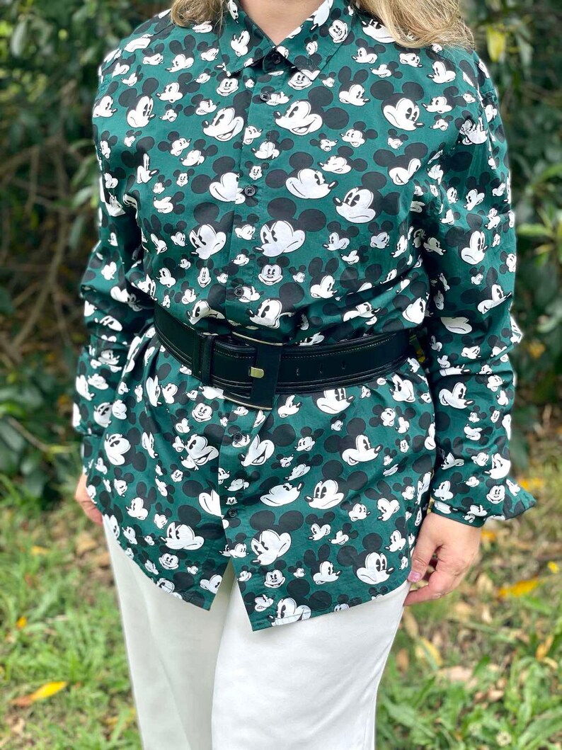 May include: A green button-down shirt with a white Mickey Mouse print all over. The shirt has long sleeves and a black leather belt.
