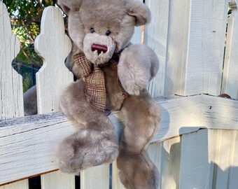 May include: A plush, light brown teddy bear with a plaid scarf, sitting on a white picket fence. The bear has a dark red nose and is made of soft, fluffy material. The bear's arms and legs are draped over the fence.