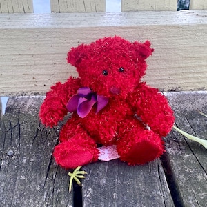May include: A red plush teddy bear with a purple bow sitting on a wooden surface.