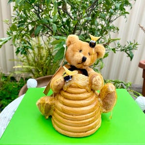 May include: A brown teddy bear figurine sitting inside a honey comb with two small yellow and black bee figurines on top of the comb.