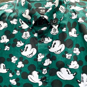 May include: A green button-down shirt with a white Mickey Mouse print all over. The shirt has a black collar and a black button placket. The label on the shirt reads "H&M Regular Fit".