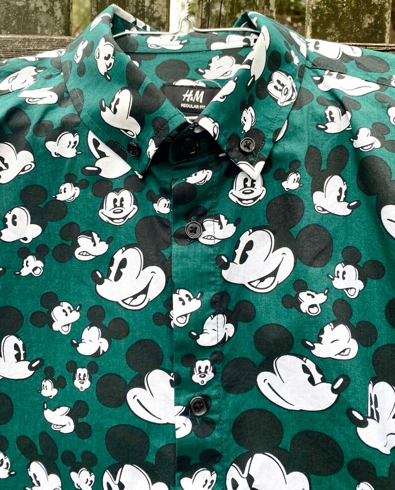 May include: A green button-down shirt with a white Mickey Mouse print all over. The shirt has a black collar and a black button placket. The label on the shirt reads "H&M Regular Fit".