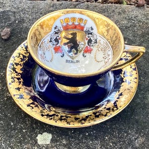 May include: A vintage teacup and saucer set. The cup and saucer are navy blue with gold trim. The inside of the cup features a crest with a bear and the word "BERLIN". The set is placed on a textured surface.