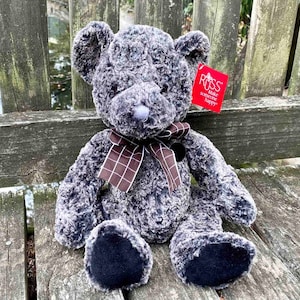 以下が含まれることがあります： 茶色と黒の格子縞のリボンが付いた灰色のぬいぐるみクマ。クマは木のベンチに座っています。クマのタグには「Russ Make Someone Happy.」と書かれています。