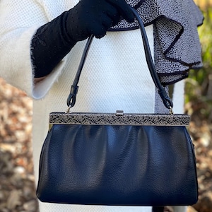 May include: A navy blue vintage handbag with a decorative metal frame and clasp. The bag has a pleated design and a structured shape. The handles are thin and black. The bag is held by a person wearing black gloves and a white coat.