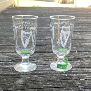 May include: Two clear glass shot glasses with a white harp design and the text "Irish Coffee Irish" on the side. The glasses are on a wooden surface.