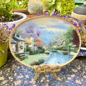 May include: A porcelain plate with a gold rim features a painting of a quaint village scene with houses, trees, and a river. The plate is on a gold metal stand.