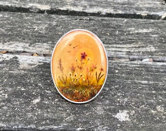 May include: Oval-shaped brooch with a silver-colored border. The brooch features a miniature landscape painting with a warm orange background, wildflowers, and three birds in flight. The artwork is encased in a clear resin.
