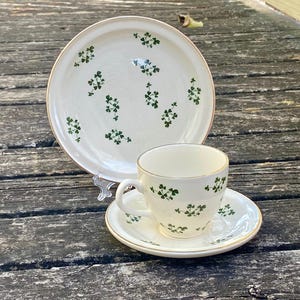 May include: A cream-colored tea set with a gold rim and green shamrock pattern. The set includes a large plate, a teacup, and a saucer. The teacup has a handle and the saucer is round. The set is on a weathered wooden surface.