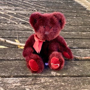May include: A burgundy teddy bear with a red bow and heart-shaped accents on its paws. The plush toy is sitting on a weathered wooden surface, with a branch and leaves in the background. The bear's fur is a deep, rich color.
