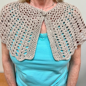 Puede incluir: Un chaleco de crochet beige con cierre de botón. El diseño calado del chaleco es visible, usado sobre una camiseta sin mangas turquesa. El chaleco es una prenda ligera y aireada.