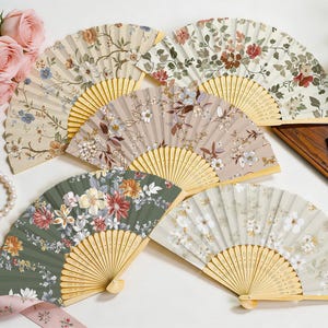 May include: Several hand fans with floral patterns in shades of green, beige, and tan. The fans have wooden handles and are displayed on a white surface. Some are open, revealing detailed designs. A spare fan is also visible.