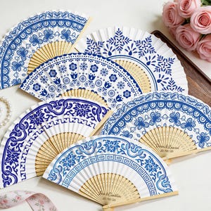 Könnte beinhalten: Eine Sammlung weißer Papierfächer mit aufwendigen blauen Blumen- und geometrischen Mustern. Die Fächer haben Holzgriffe und sind halbkreisförmig angeordnet. Einige Fächer haben personalisierten Text. Eine Perlenkette und rosa Rosen sind im Hintergrund.