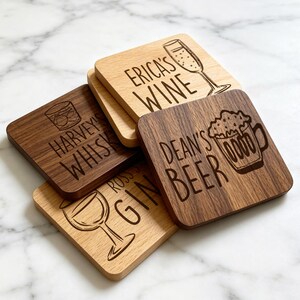 May include: Four square wooden coasters with rounded corners, featuring laser-engraved illustrations and text. The coasters display the phrases Erica's Wine, Harvey's Whiskey, Dean's Beer, and Ross's Gin, each accompanied by a matching drink icon.