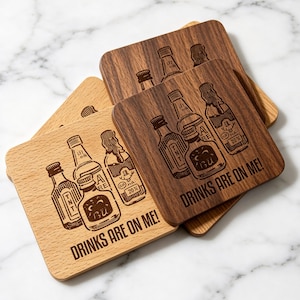 May include: A set of square wooden coasters in light and dark wood tones. Each coaster features a laser-engraved illustration of three glass bottles and the text "DRINKS ARE ON ME!" The design includes detailed bottle caps and labels.