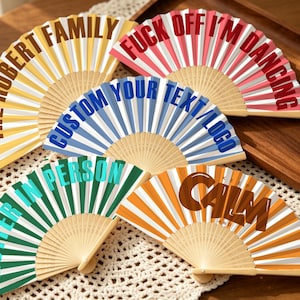 May include: Five folding hand fans with bamboo ribs and pleated fabric in yellow, pink, blue, green, and orange. The fans feature bold text including THE ROBERT FAMILY, FUCK OFF I'M DANCING, CUSTOM YOUR TEXT/LOGO, HOTTER IN PERSON, and KEEP CALM AND.