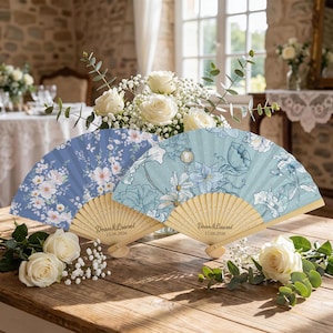May include: Two folding hand fans with light wood ribs, each featuring the text "Dean & Laurel 15.06.2026". The left fan has a dark blue background with white and pink floral patterns, while the right fan displays a light blue botanical illustration.