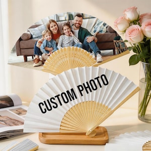 May include: White and beige folding fans with the text "CUSTOM PHOTO" printed in black. One fan is displayed on a wooden tray. A family photo is displayed on a fan in the background. A vase of pink roses is also in the background.
