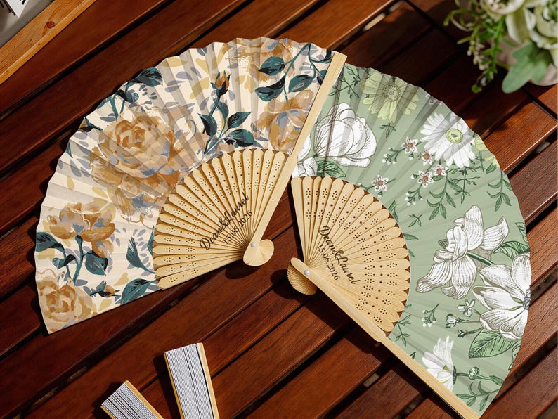 May include: Two folding hand fans with light wood frames and fabric leaves. One fan features a cream background with yellow and teal floral patterns. The other has a sage green background with white line-art flowers. Both are engraved with Dean & Laurel 15.06.2026.
